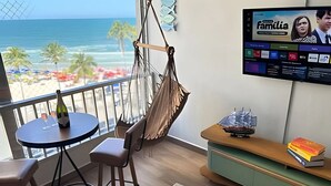 Un balcón con vistas a la playa, una hamaca, una mesa con una botella y un televisor mostrando un programa.