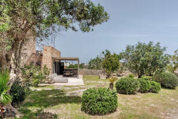 A stone building with a covered outdoor seating area, surrounded by well-maintained greenery and a clear sky.