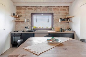 A compact kitchen with a large window, wooden shelves, and a dining area with a table.
