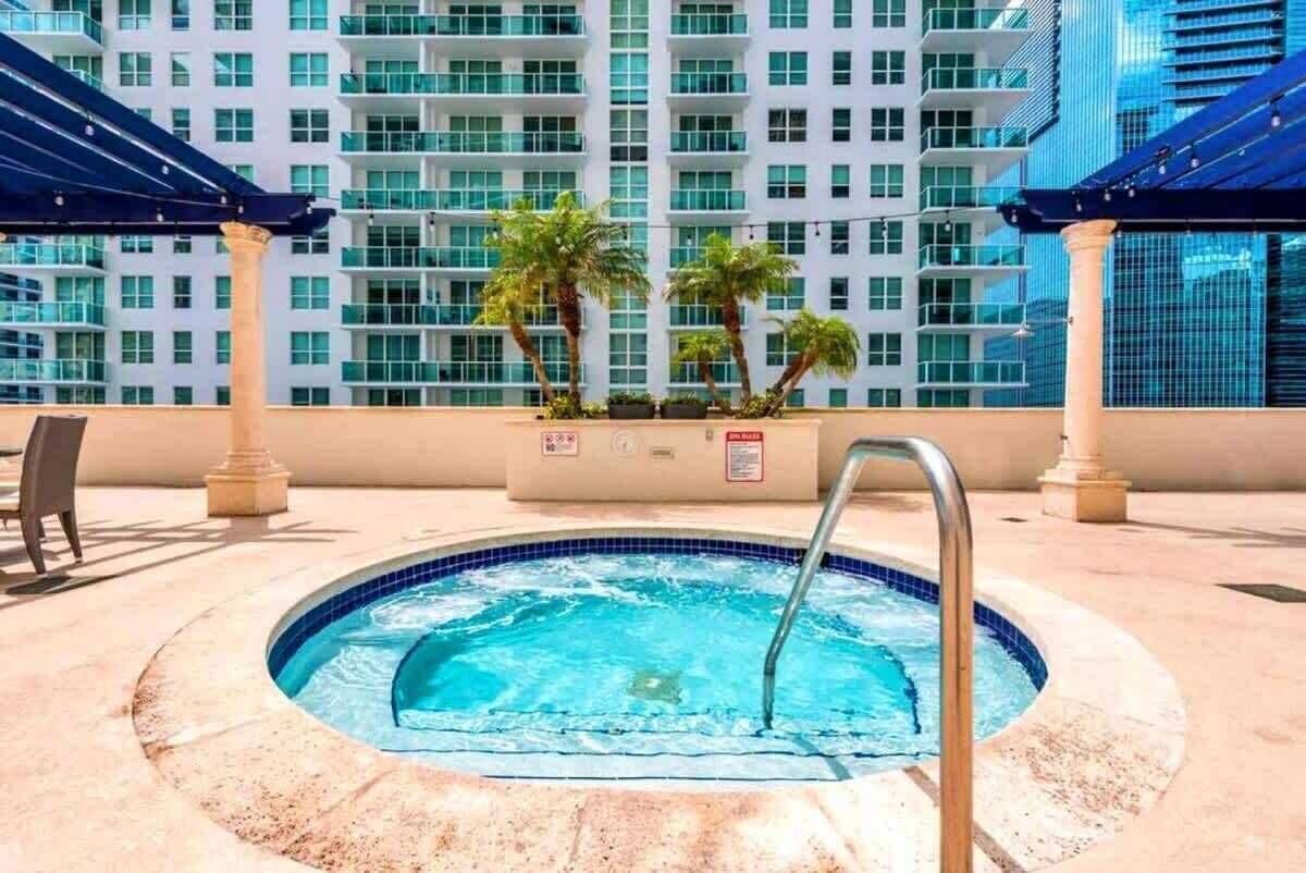 A rooftop pool with a blue tiled edge, surrounded by beige pillars and palm trees, with a modern building in the background.