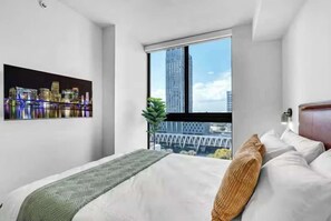 A hotel room with a large bed, a cityscape view through a window, and a framed picture on the wall. - 4BR/4BA 2-Unit Combo for 12 · Rooftop Pool + Views (Miami)