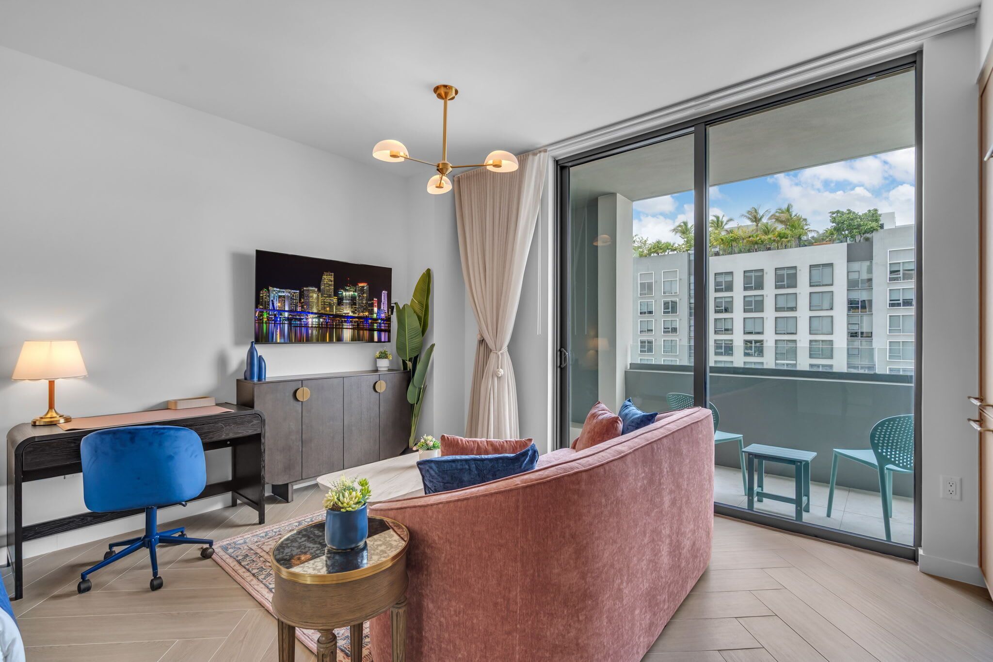 Un salon moderne comprenant un bureau, un canapé, un téléviseur et un balcon avec vue sur les immeubles.