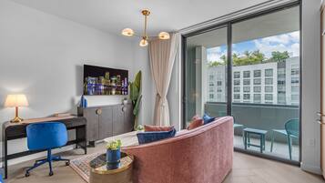 Un salon moderne comprenant un bureau, un canapé, un téléviseur et un balcon avec vue sur les immeubles.