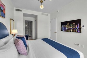 Una camera d'albergo con un letto, un comodino, una lampada, un quadro con una vista cittadina e una porta che dà su un'altra stanza.