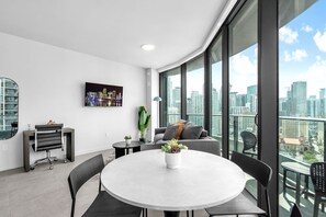 A modern office space with a round table, chairs, a desk with a chair, a potted plant, and a city view through large windows. - 2 Apts for 8 · 2BR/2BA, Rooftop Pool, Downtown MIA (Miami)