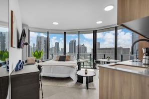 A modern hotel room with a bed, a small dining area, a kitchen, and large windows offering a city view. - 2 Apts for 8 · 2BR/2BA, Rooftop Pool, Downtown MIA (Miami)