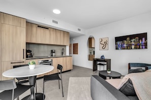 A modern kitchen with wooden cabinets, a dining area with a round table and chairs, a sofa, and a cityscape painting on the wall. - 2 Apts for 8 · 2BR/2BA, Rooftop Pool, Downtown MIA (Miami)