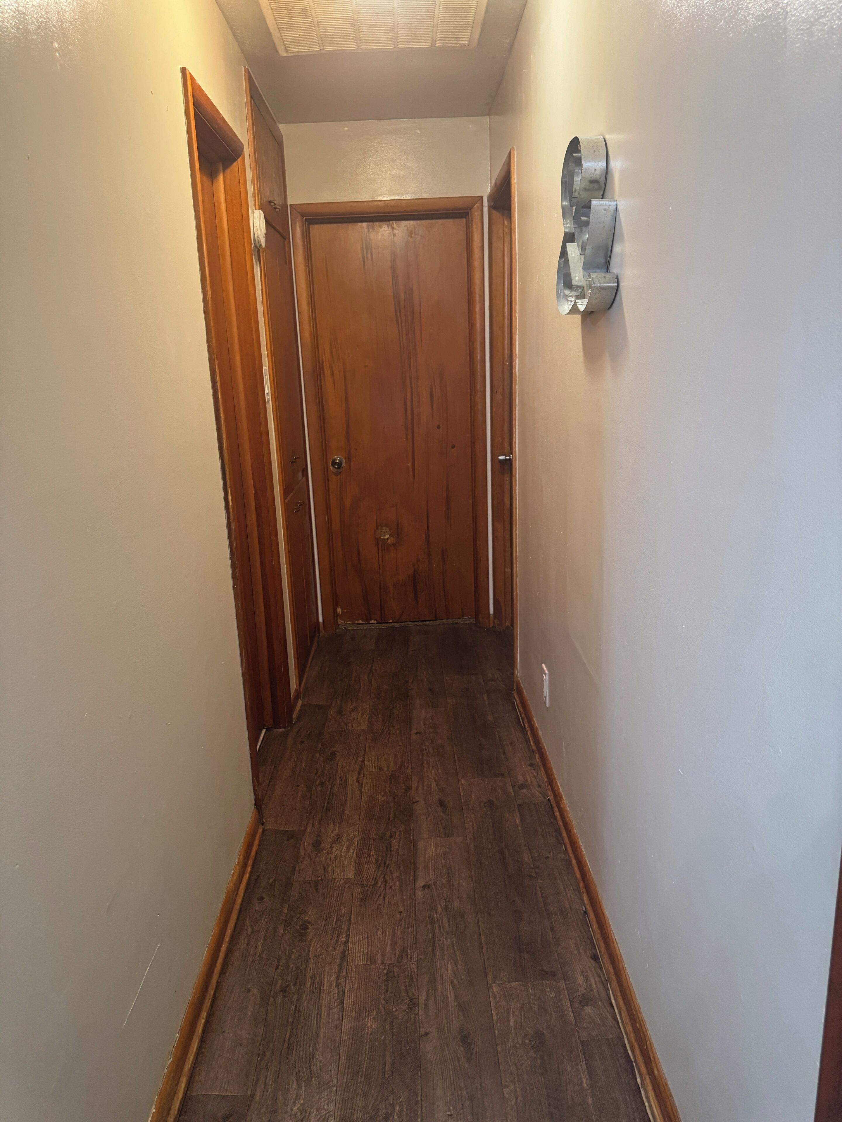 A narrow hallway with wooden flooring, a wooden door at the end, and a wall-mounted light fixture.