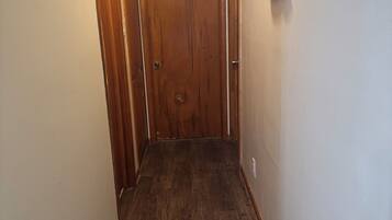 Un couloir étroit au plancher de bois, une porte en bois au fond et un luminaire fixé au mur.