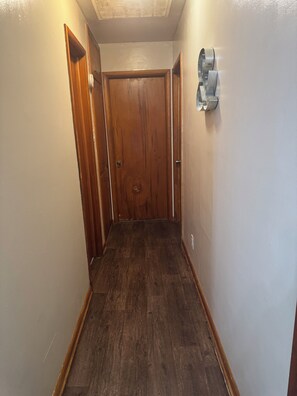 A narrow hallway with wooden flooring, a wooden door at the end, and a wall-mounted light fixture.