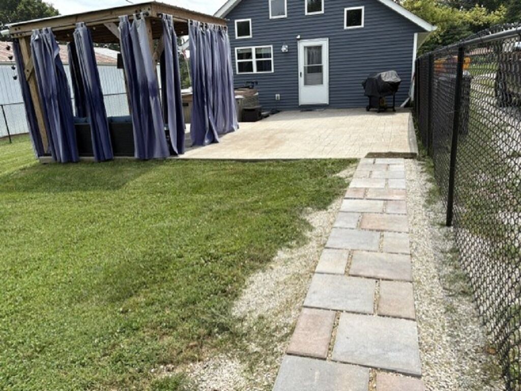 A backyard with a patio, a covered area with curtains, a house, and a chain-link fence.