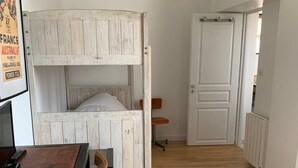 A room with a bunk bed, a desk with a chair, a hanging light fixture, and a door leading to another room.