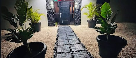 A pathway leading to a building entrance with a 'OPEN' sign, flanked by potted plants.