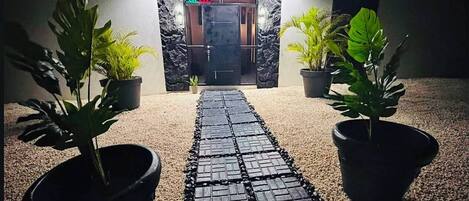 Un camino que lleva a la entrada de un edificio, con una señal que dice 'OPEN', flanqueado por macetas con plantas.