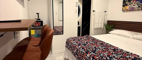 A hotel room with a bed, a desk with a chair, a TV mounted on the wall, a mirror, and a painting.