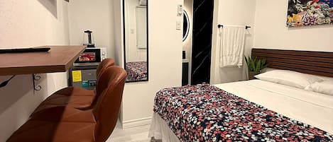 A hotel room with a bed, a desk with a chair, a TV mounted on the wall, a mirror, and a painting.