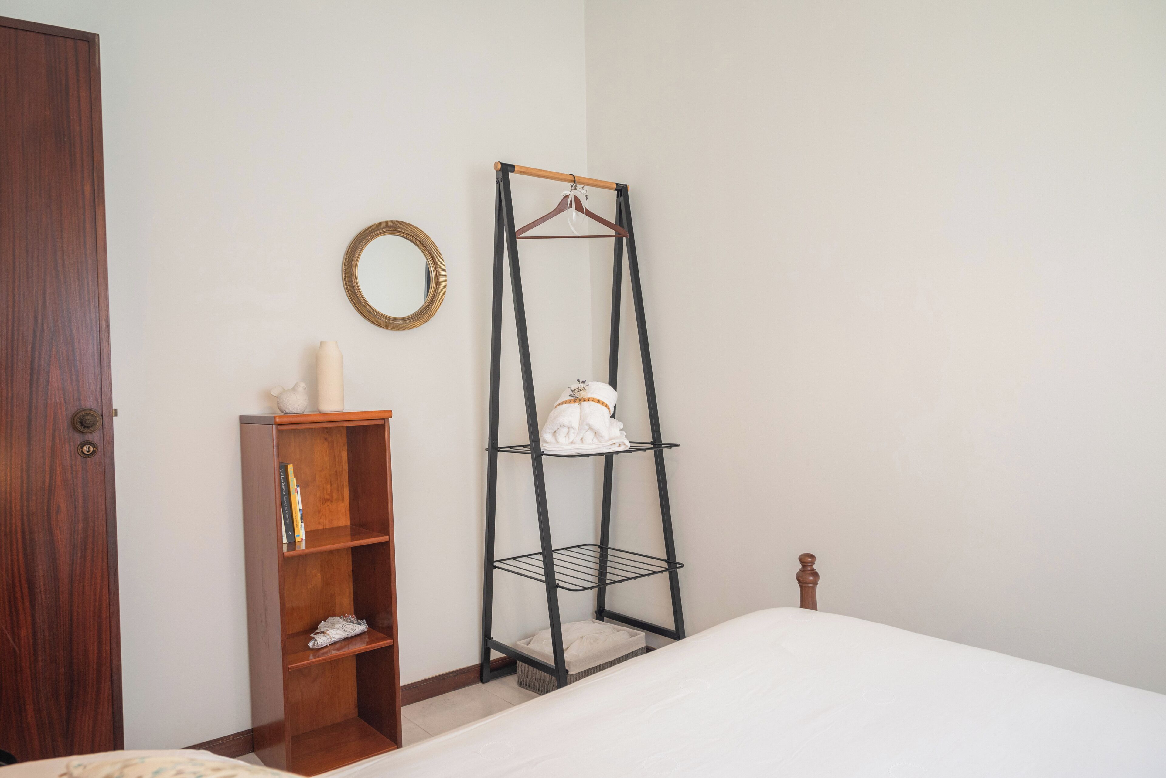 Une chambre avec une armoire en bois, une étagère en métal noir, un lit blanc et un miroir rond accroché au mur.