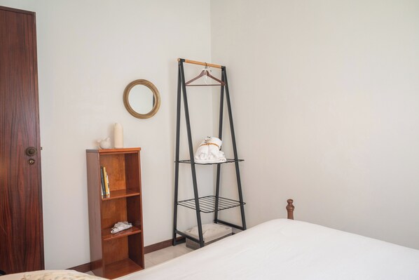 Une chambre avec une armoire en bois, une étagère en métal noir, un lit blanc et un miroir rond accroché au mur.