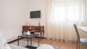 Un salón con televisión de pantalla plana montada en la pared, un escritorio de madera con un florero y una planta pequeña, una mesa de comedor con una silla, y cortinas de encaje en la ventana.