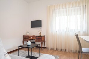 A living room with a flat-screen TV mounted on the wall, a wooden desk with a vase and a small plant, a dining table with a chair, and sheer curtains on the window. - Memories - Local Lodging (Nogueira da Regedoura)