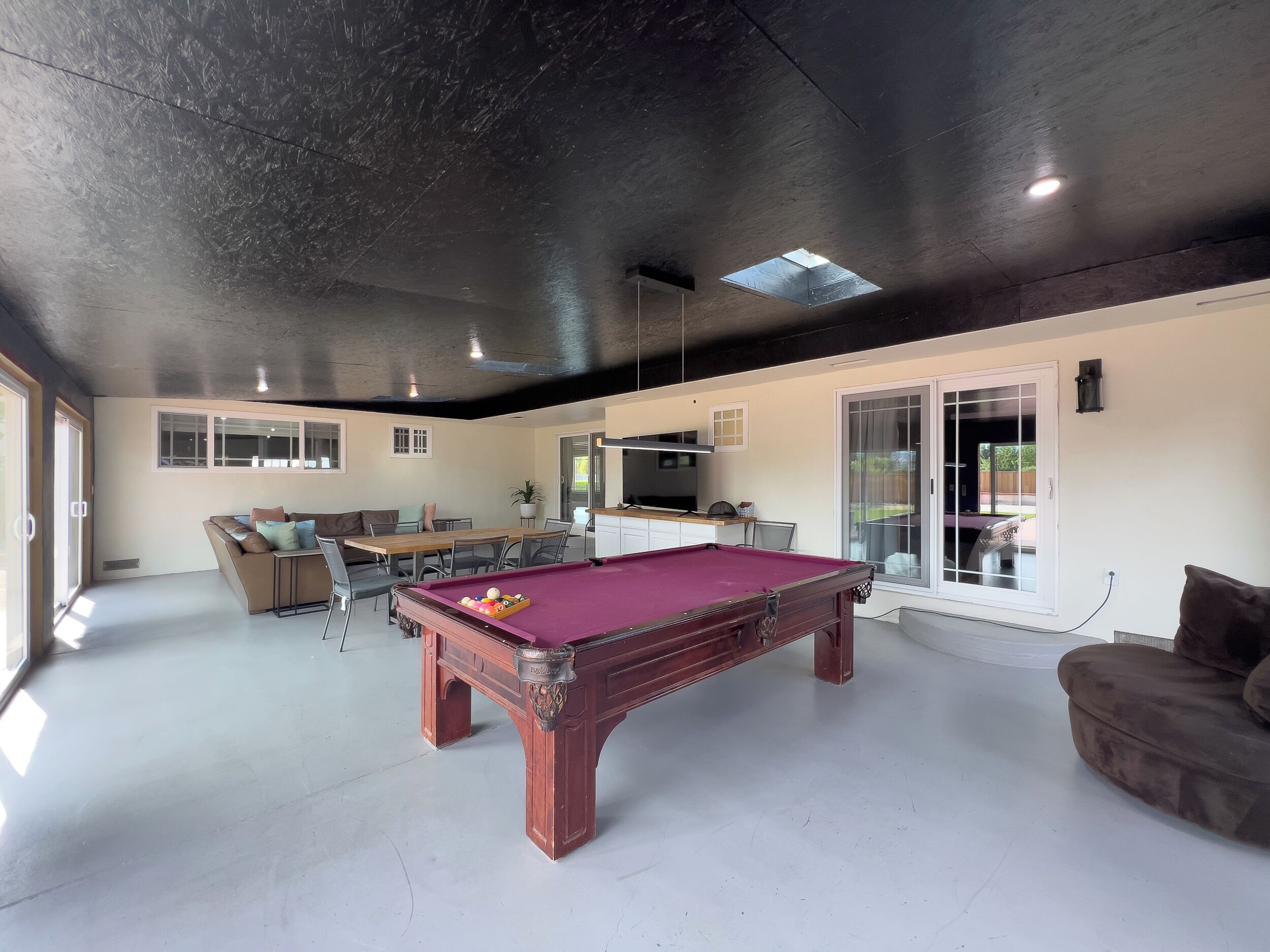 A spacious indoor area with a pool table, a sofa, a dining table, and a kitchen area.