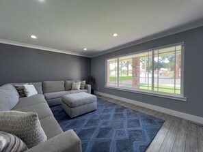 A modern living room with a sectional sofa, a blue patterned rug, and a large window with a view of a playground. - Beautiful 4-bedroom house with AC in Hollister (Hollister)
