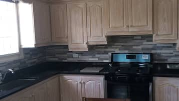Una cocina con alacenas de madera clara, encimera negra, estufa de acero inoxidable y una mesa de comedor con una silla.