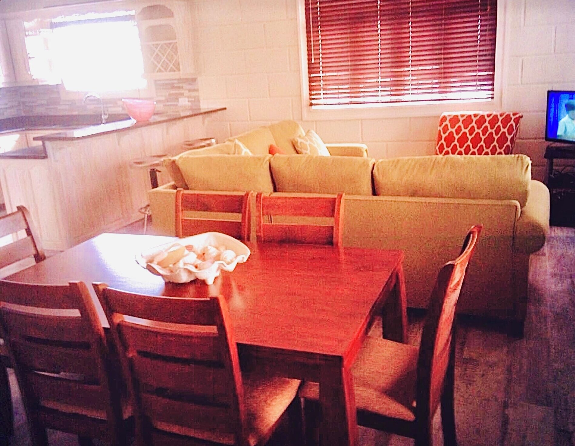A living room with a sofa set, a dining table with chairs, a television, and a kitchen area with a window.