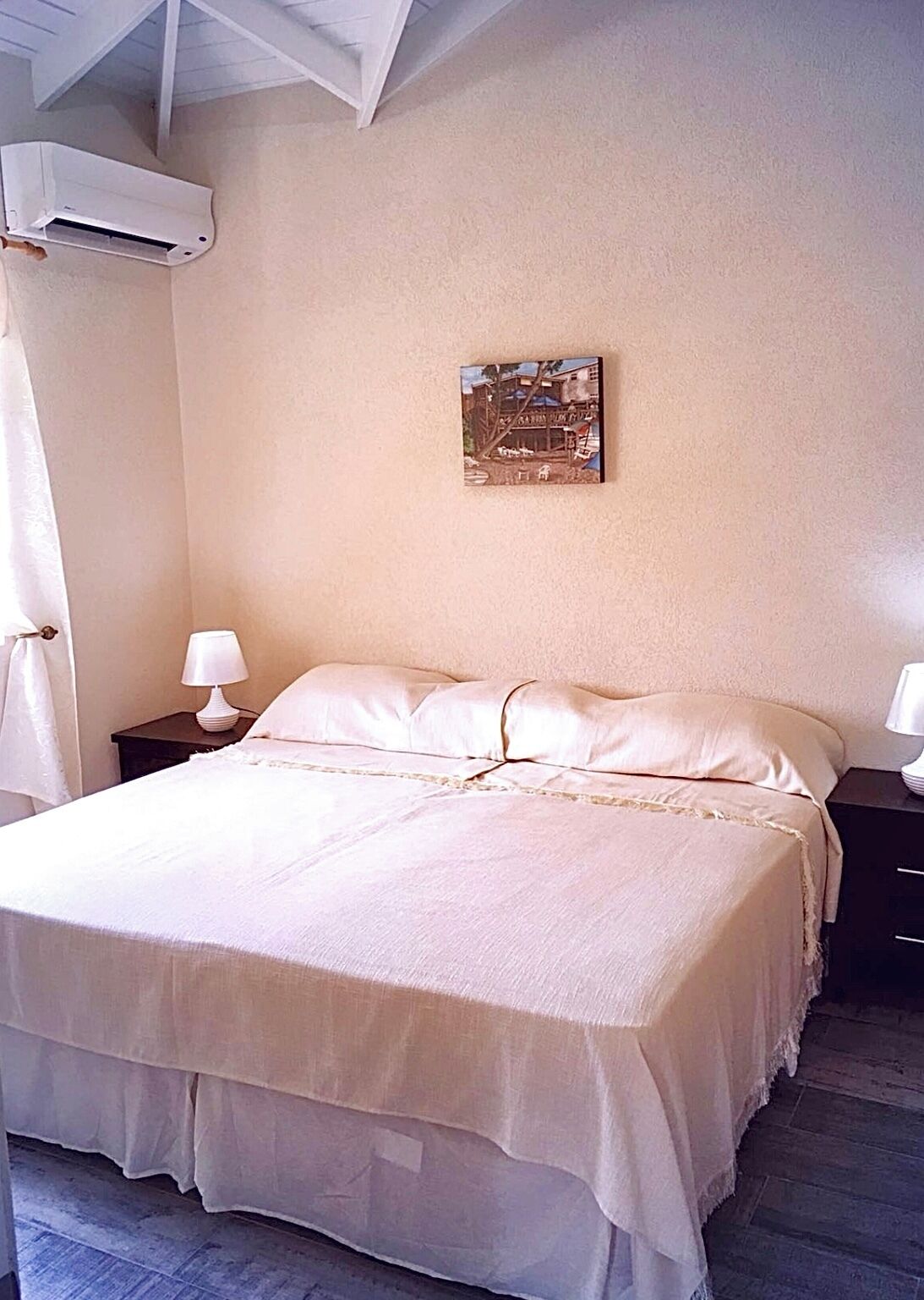 Un dormitorio con cama, dos mesitas de noche con lámparas, aire acondicionado en la pared y un cuadro enmarcado.