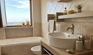 A bathroom with a bathtub, a sink, and a window with a view of the sea. - Romantic Attic Room with Ocean View for Couples (Porto Torres)