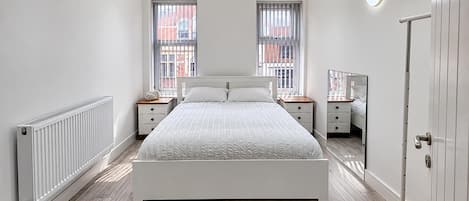 Ein Schlafzimmer mit einem weißen Bett, zwei Nachttischen, einer Heizung und einem großen Fenster mit Jalousien.
