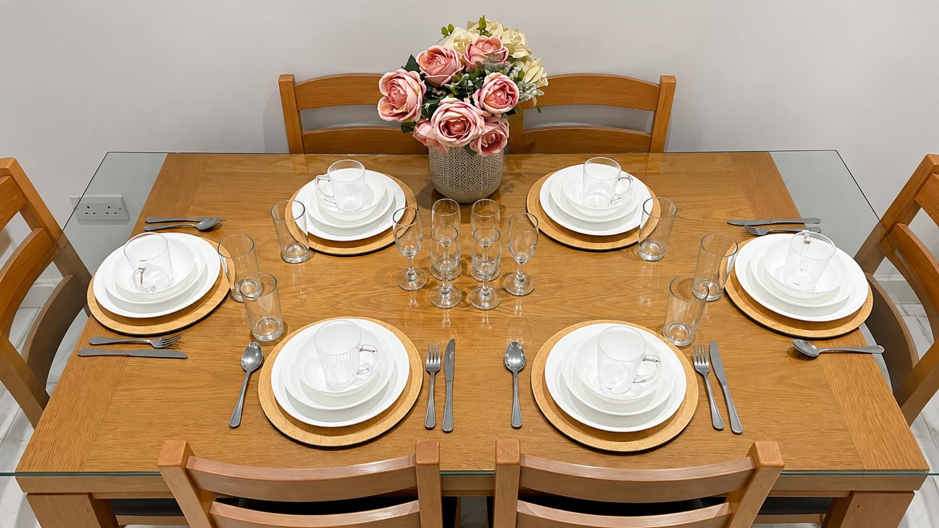 Une table à manger est dressée pour six personnes, avec de l’argenterie blanche, des verres à bord doré et des couverts. Un vase rempli de fleurs roses et blanches est placé au centre de la table.