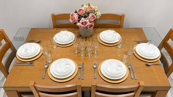 Une table à manger est dressée pour six personnes, avec de l’argenterie blanche, des verres à bord doré et des couverts. Un vase rempli de fleurs roses et blanches est placé au centre de la table.