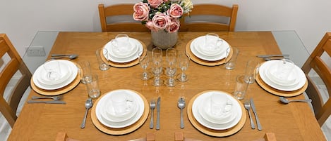 Mesa de jantar posta para seis com pratos brancos, copos com detalhes dourados e talheres. Um vaso com flores rosa e branca está centralizado na mesa.