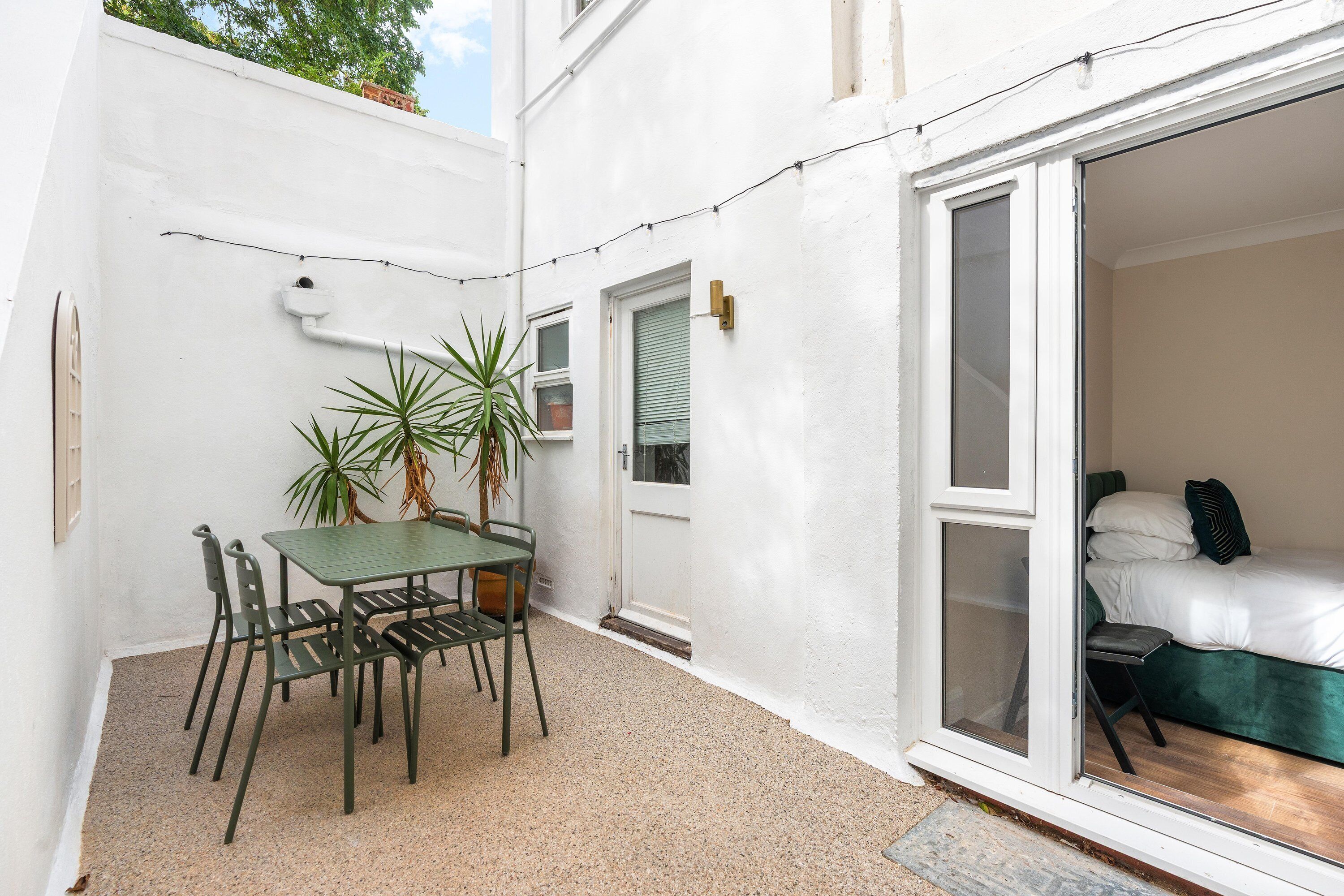 A small outdoor patio with a table and chairs, a potted plant, and a sliding glass door leading to a bedroom with a bed and pillows.