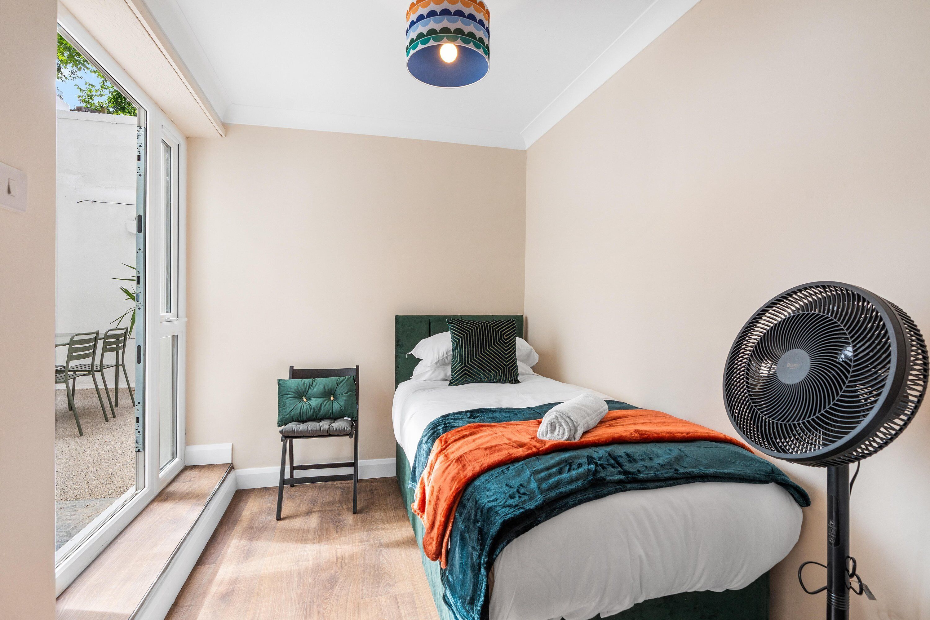 Ein Schlafzimmer mit einem Bett, einem Sessel, einem Standventilator und einem Fenster.