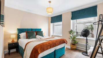 Een slaapkamer met een bed, nachtkastje, lamp, raam met gordijnen, een kamerplant, radiator en een trap.