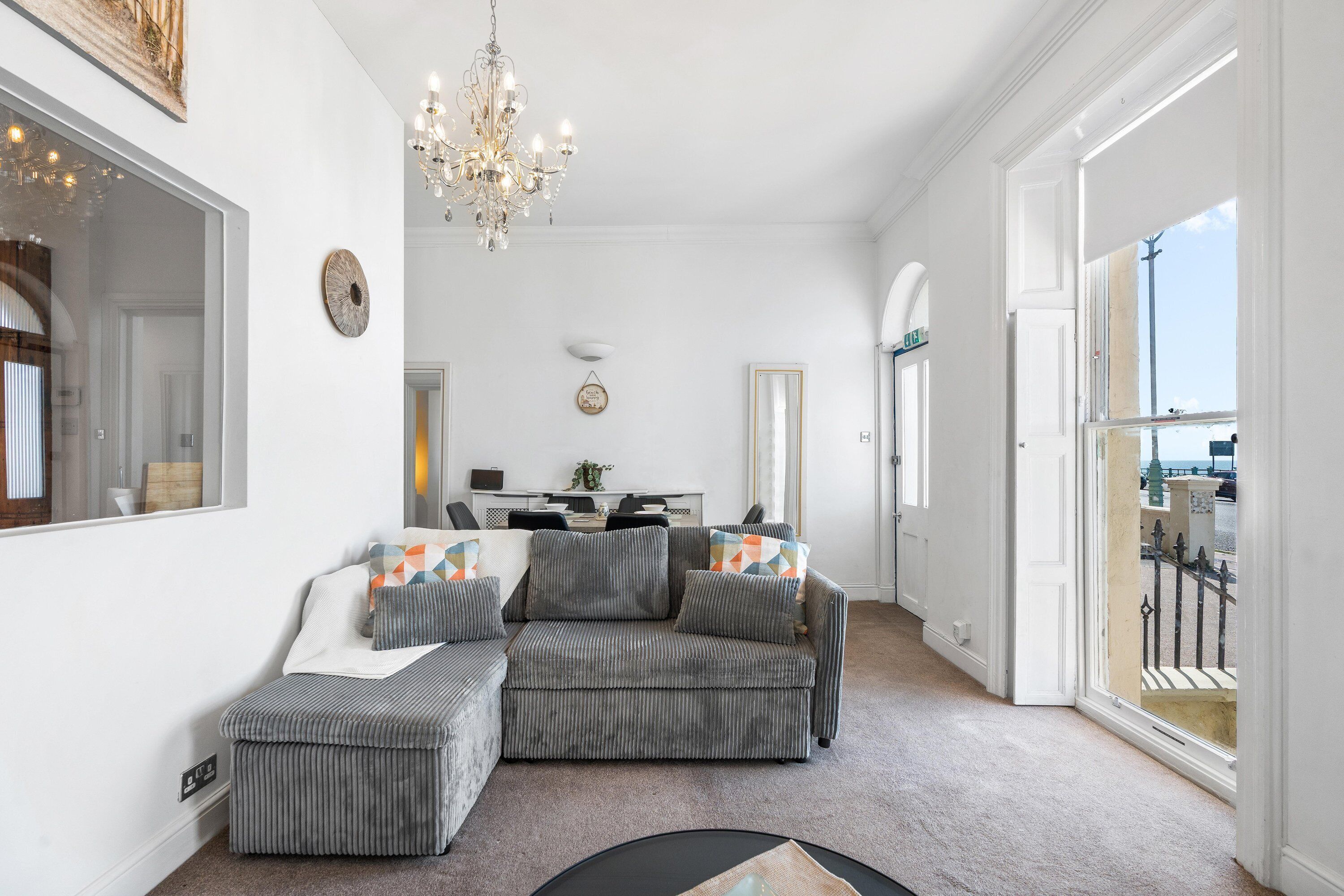 A living room with a grey sofa, ottoman, chandelier, and a view through a large window.
