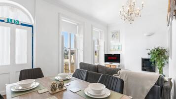 A modern dining area with a table set for two, a sofa, and a television displaying 'NETFLIX' in the background.