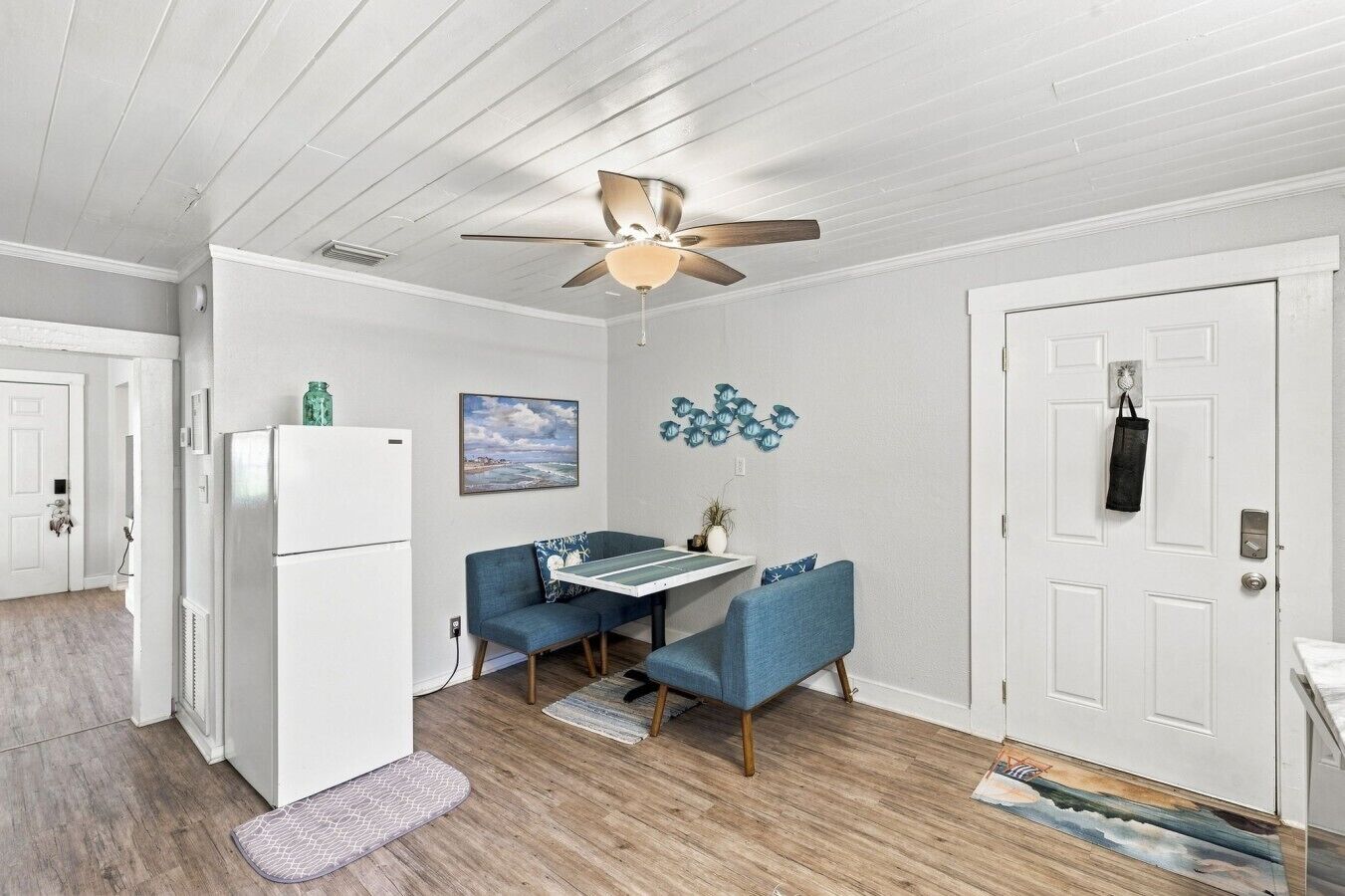 A compact kitchen with a white refrigerator, a dining table with blue chairs, a ceiling fan, and a door with a coat hanging on it.