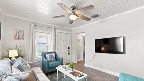 Uma sala de estar com ventilador de teto, televisão fixada na parede, sofá, mesa de centro e uma cadeira.