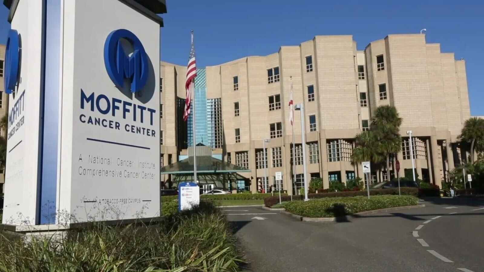 Rakennuksessa on Moffitt Cancer Center -keskuksen kyltti, ja siinä on selkeä sisäänkäynti, pysäköintialue sekä lipputanko, jossa liehuu Yhdysvaltain lippu.