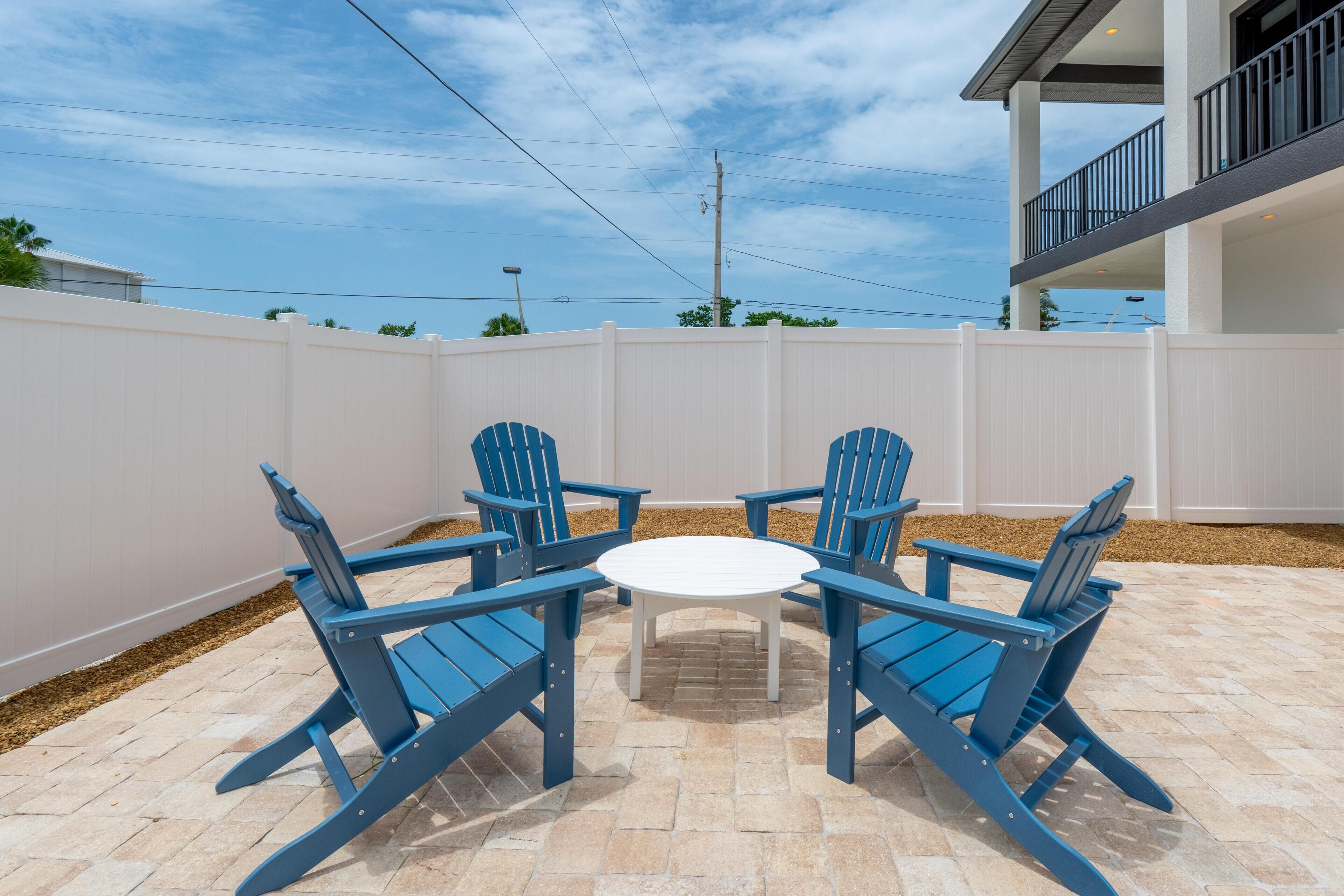 A patio with four blue Adirondack chairs and a white table, surrounded by a white fence.