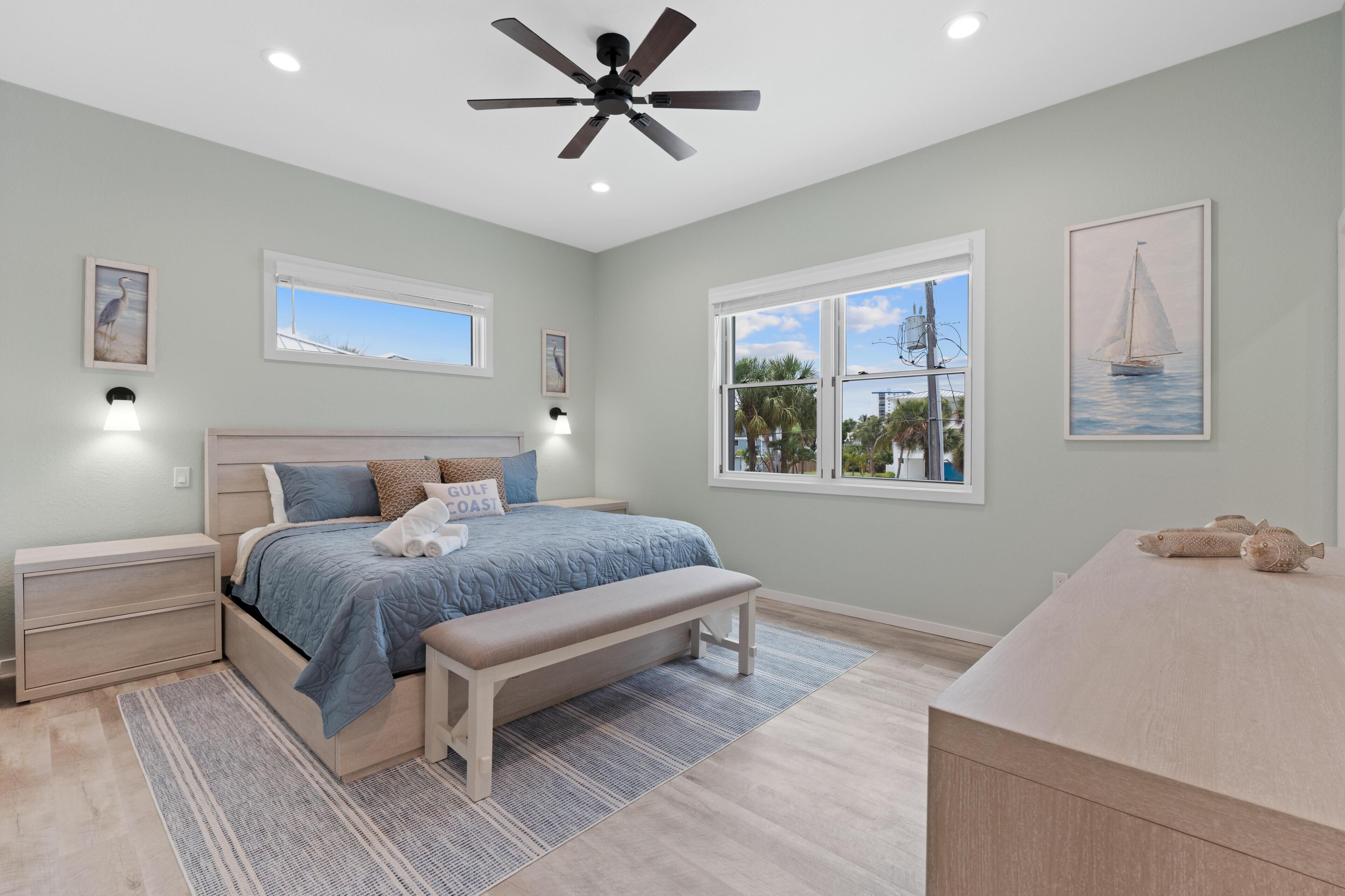 A bedroom with a bed, bedside tables, a bench, a ceiling fan, and artwork on the walls.