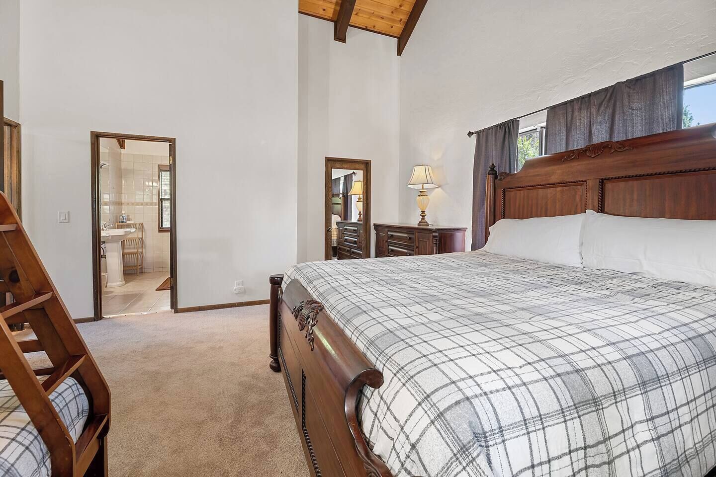 Un dormitorio con una cama grande, cabecera de madera, una mesita de noche con lámpara, un aparador y un espejo. Se ve una silla de madera en la esquina.
