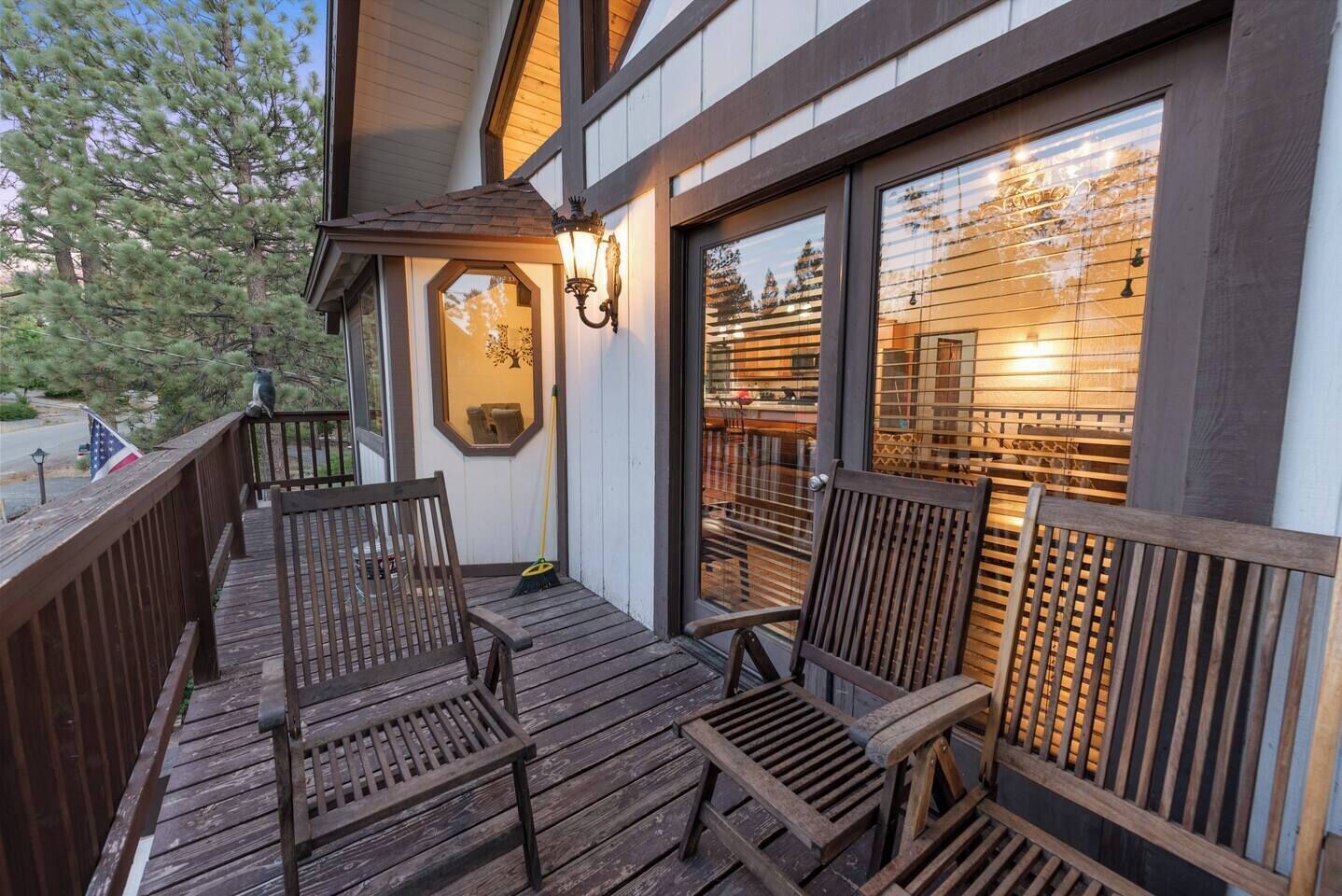 A wooden deck with two chairs and a small table, a window with a view of trees, and a wall-mounted light fixture.