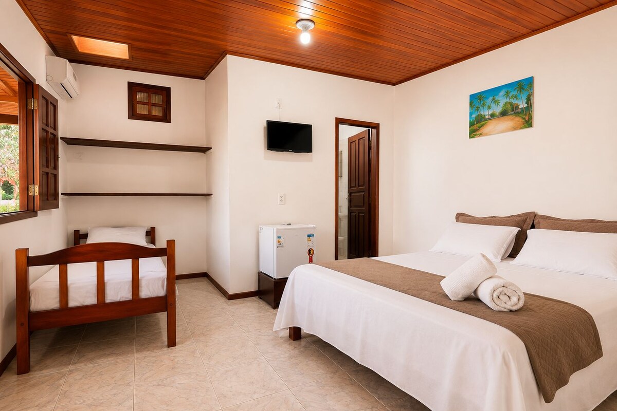 Un dormitorio con cama, una silla de madera, televisor, refrigerador y un cuadro en la pared.