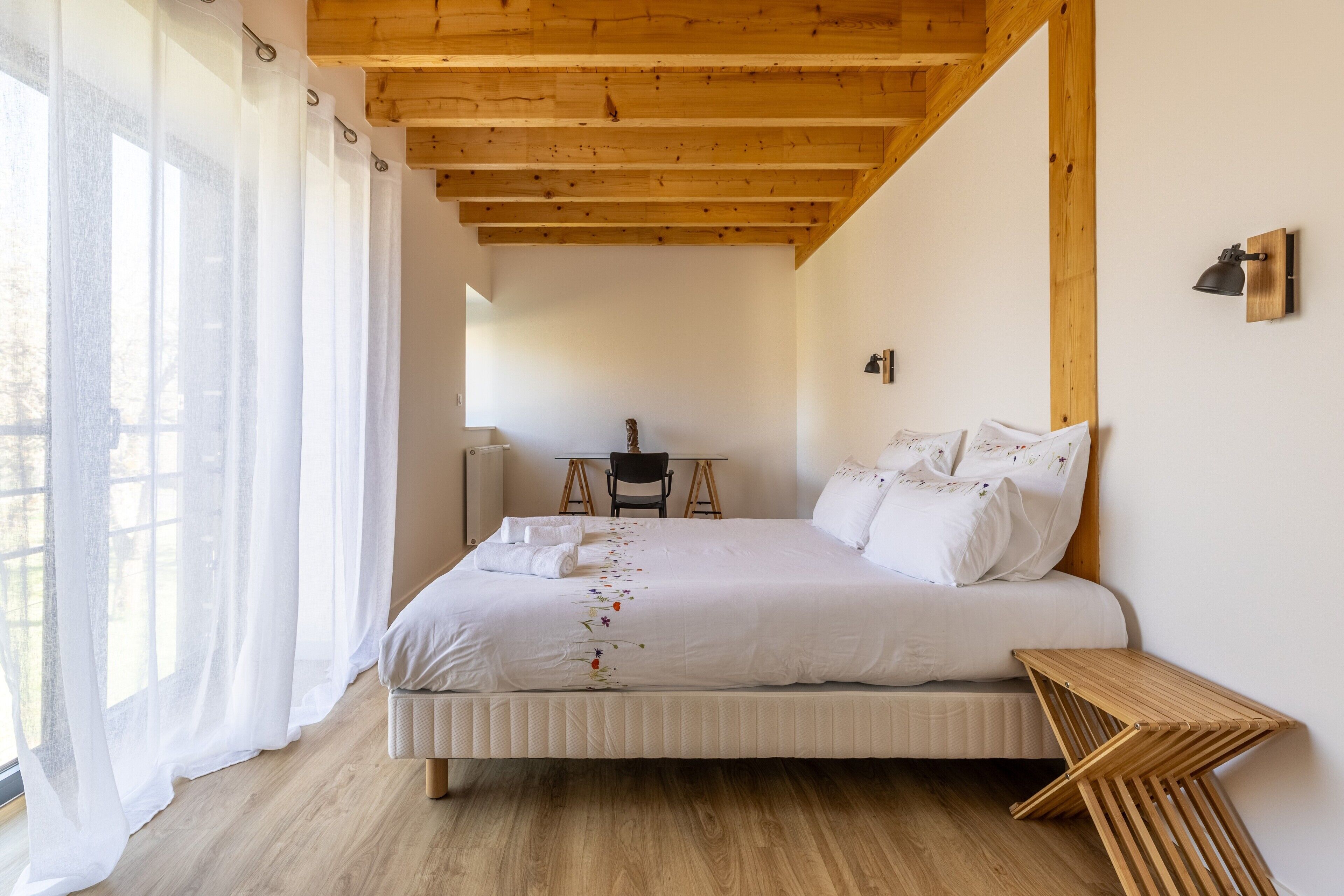 Un dormitorio con techo de madera, una cama con ropa de cama blanca, una mesita y una silla.