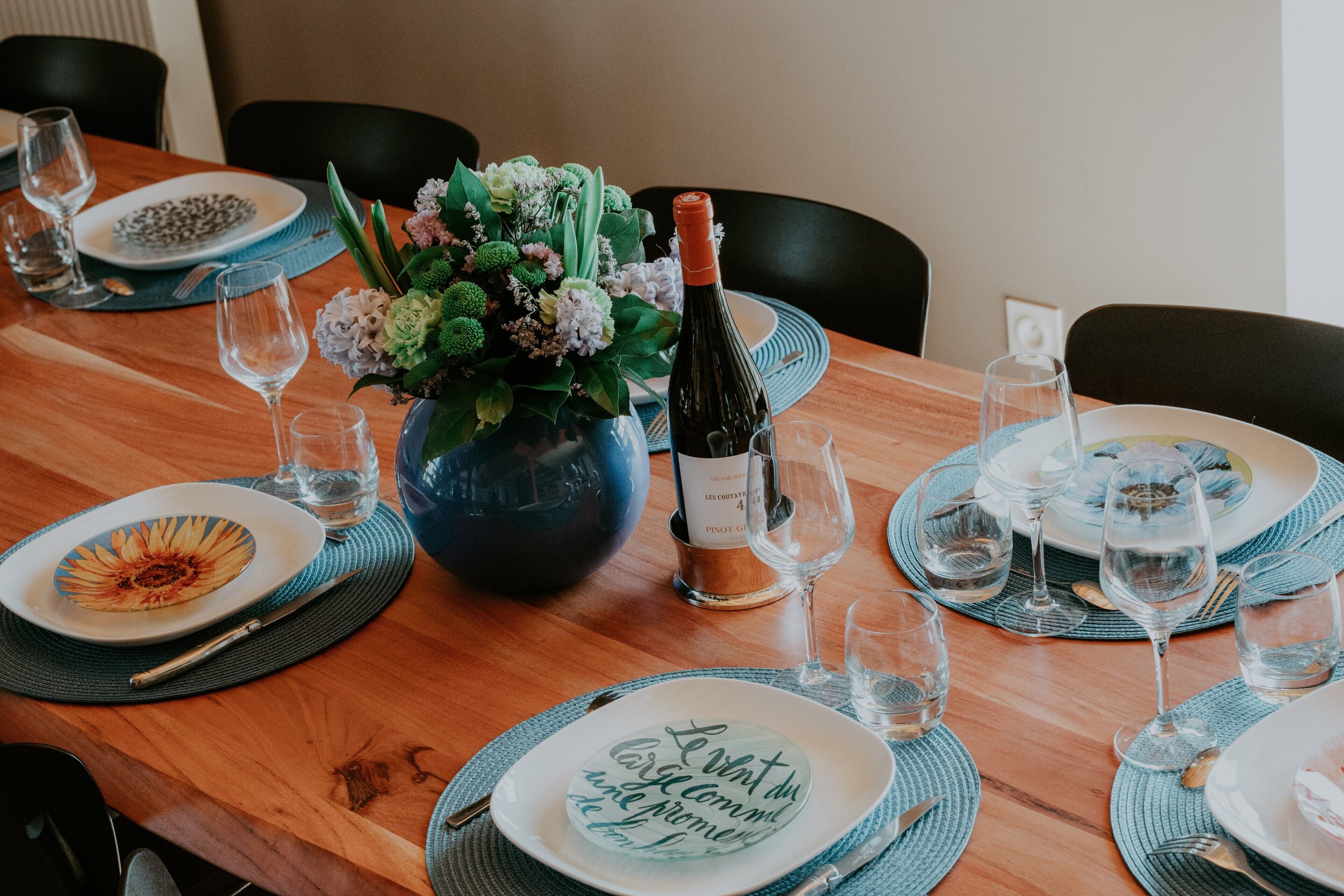 Une table à manger dressée avec des assiettes, des verres, une bouteille de vin et un vase de fleurs.