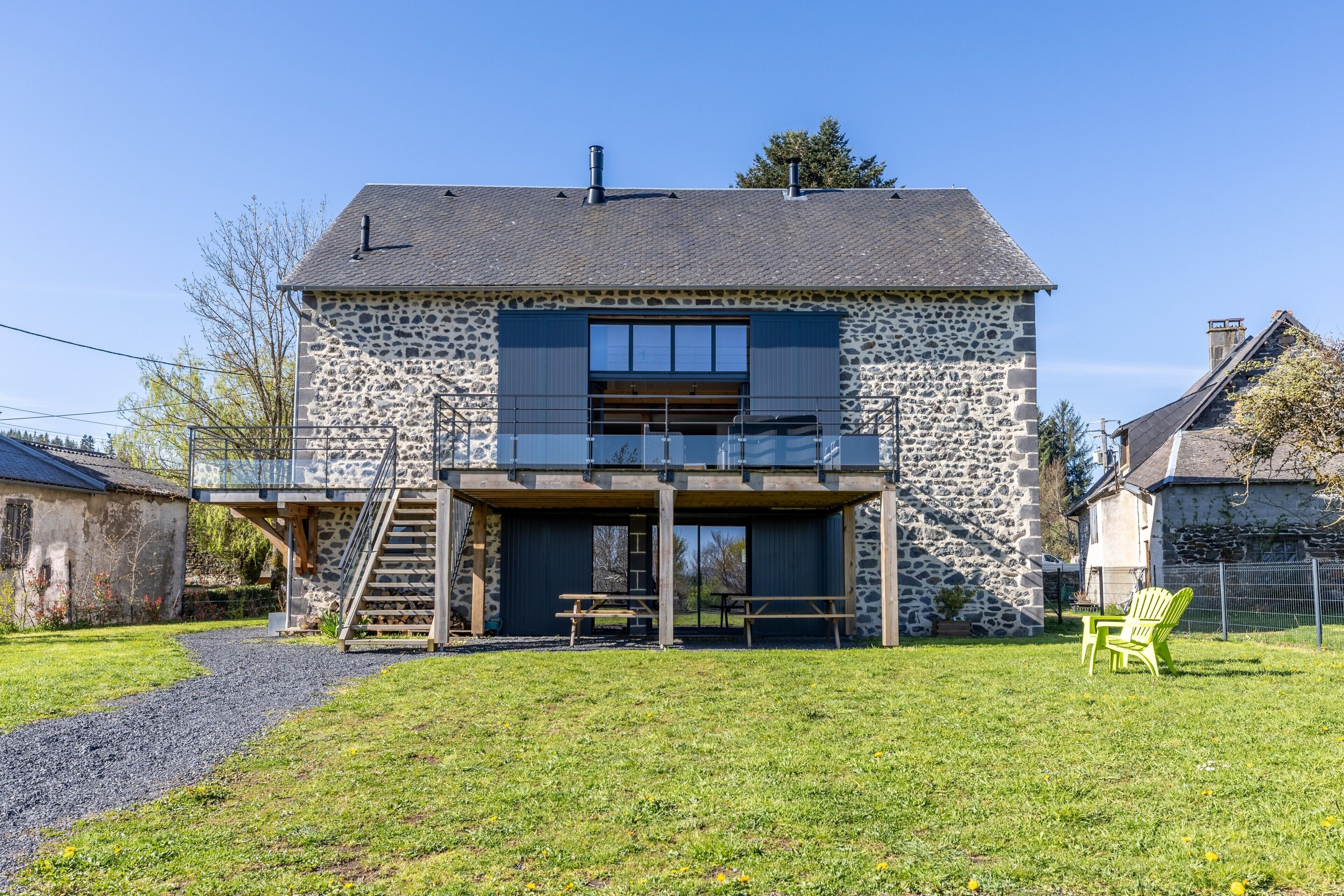 Une maison en pierre à deux étages, dotée d’une terrasse en bois, d’un escalier et d’un espace extérieur aménagé et couvert.